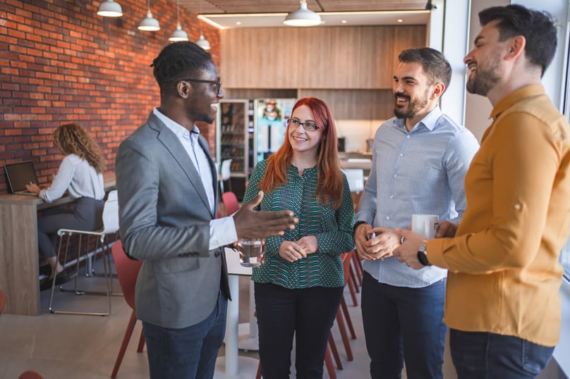 A group of coworkers laughing and having a lively conversation in a modern office break area with a brick wall and cozy ambience.