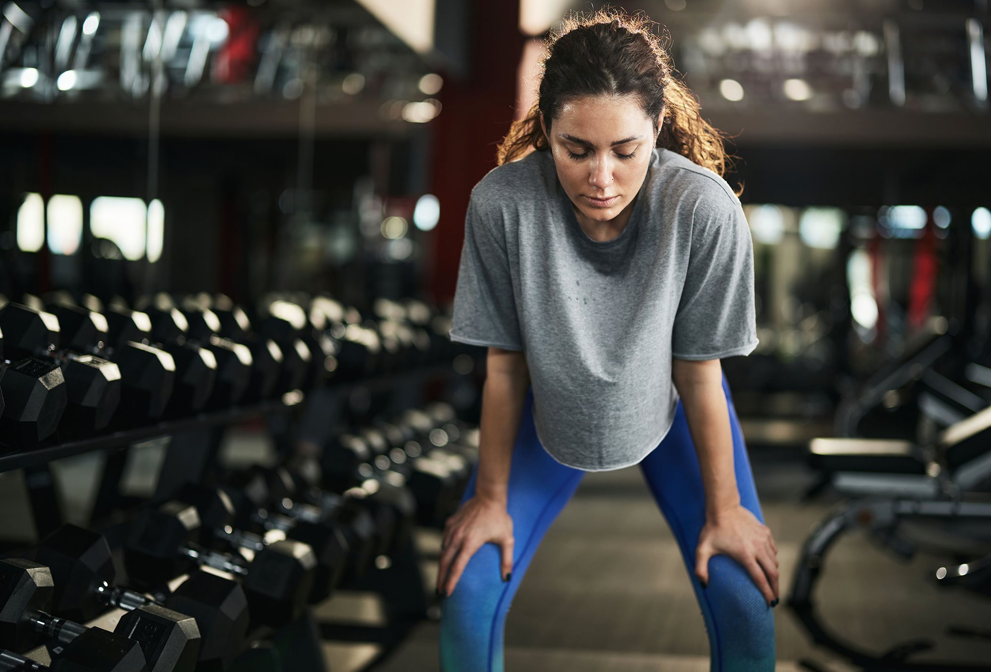 A woman in a gym leans forward, catching her breath after an intense workout, surrounded by rows of dumbbells.