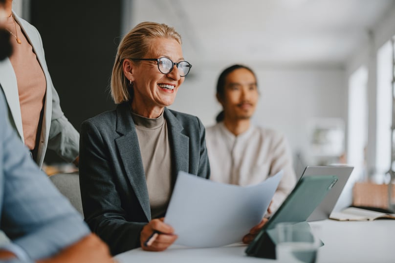 Smiling businesswoman in a meeting, holding documents and engaged in discussion with colleagues in a bright office setting.