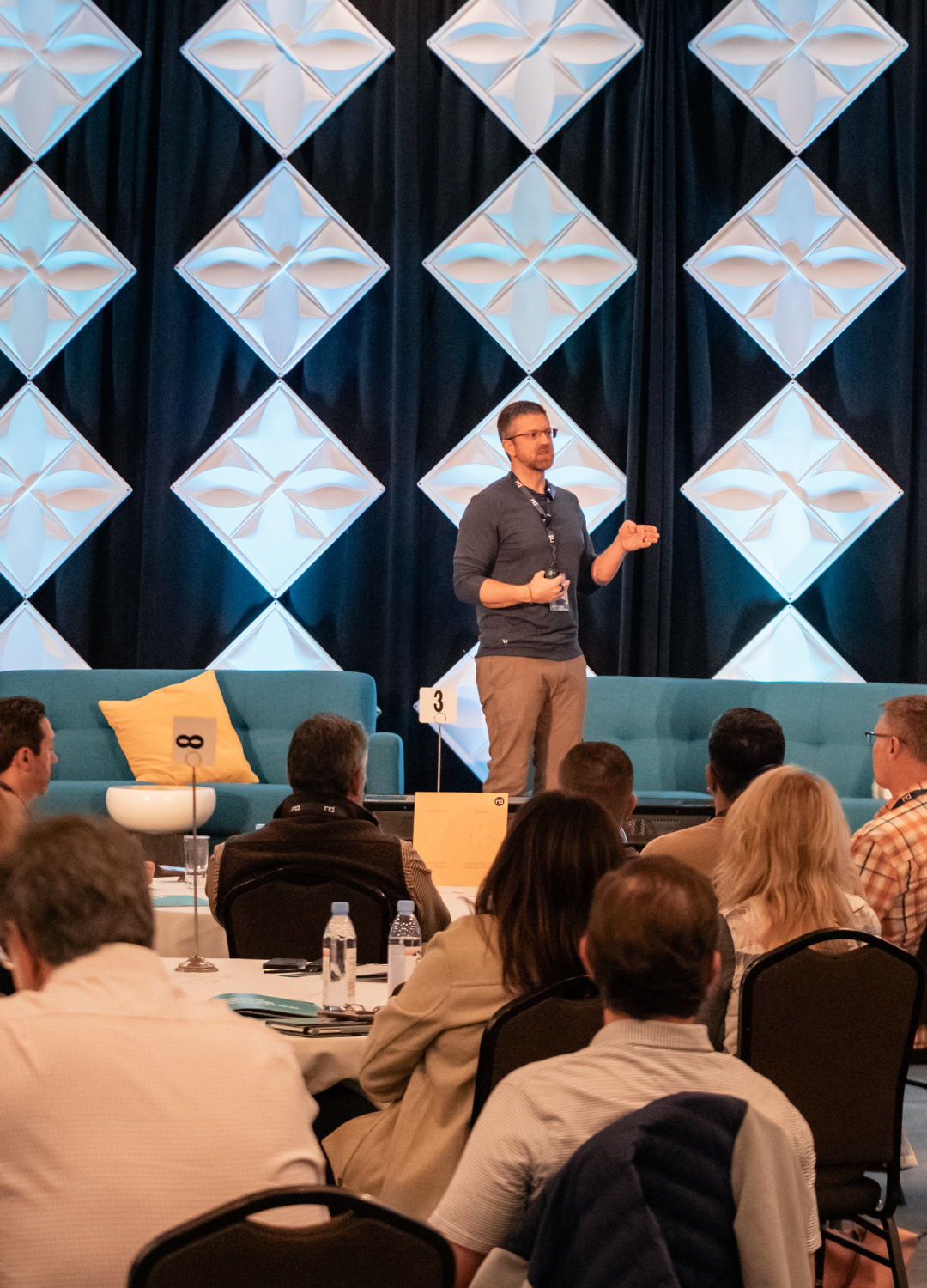 Man speaking on stage to an audience at a conference, with a geometric blue and white backdrop and a casual, professional setting.