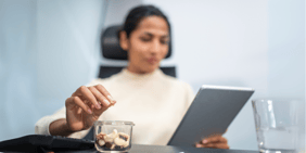 Woman sitting at a desk, focused on a tablet in one hand while picking mixed nuts from a container with the other.