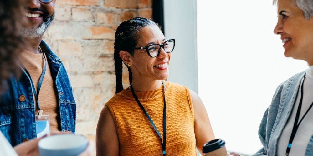 Three professionals smile and chat while holding coffee cups during a casual workplace gathering.