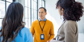  Man in a yellow sweater engaged in a focused discussion with two colleagues in a bright, modern workspace with large windows.