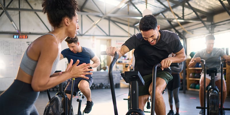 People working out on air bikes in an office gym while a coach encourages them.