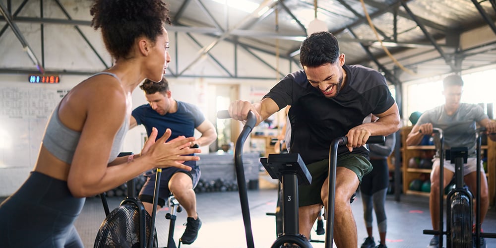 People working out on air bikes in an office gym while a coach encourages them.