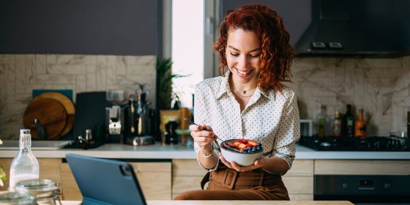 A woman with curly red hair smiles while eating from a bowl in a modern kitchen, with a laptop open on the counter.