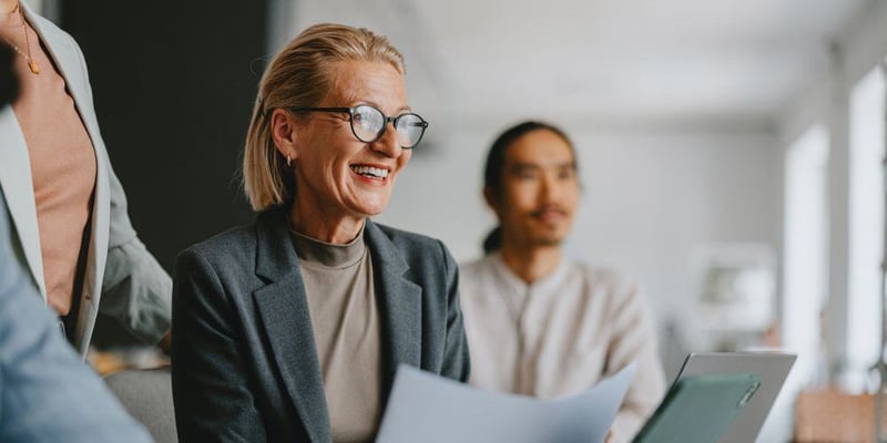 Smiling businesswoman in a meeting, holding documents and engaged in discussion with colleagues in a bright office setting.