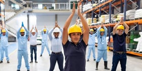 Warehouse workers in safety helmets and uniforms performing group stretching exercises together.