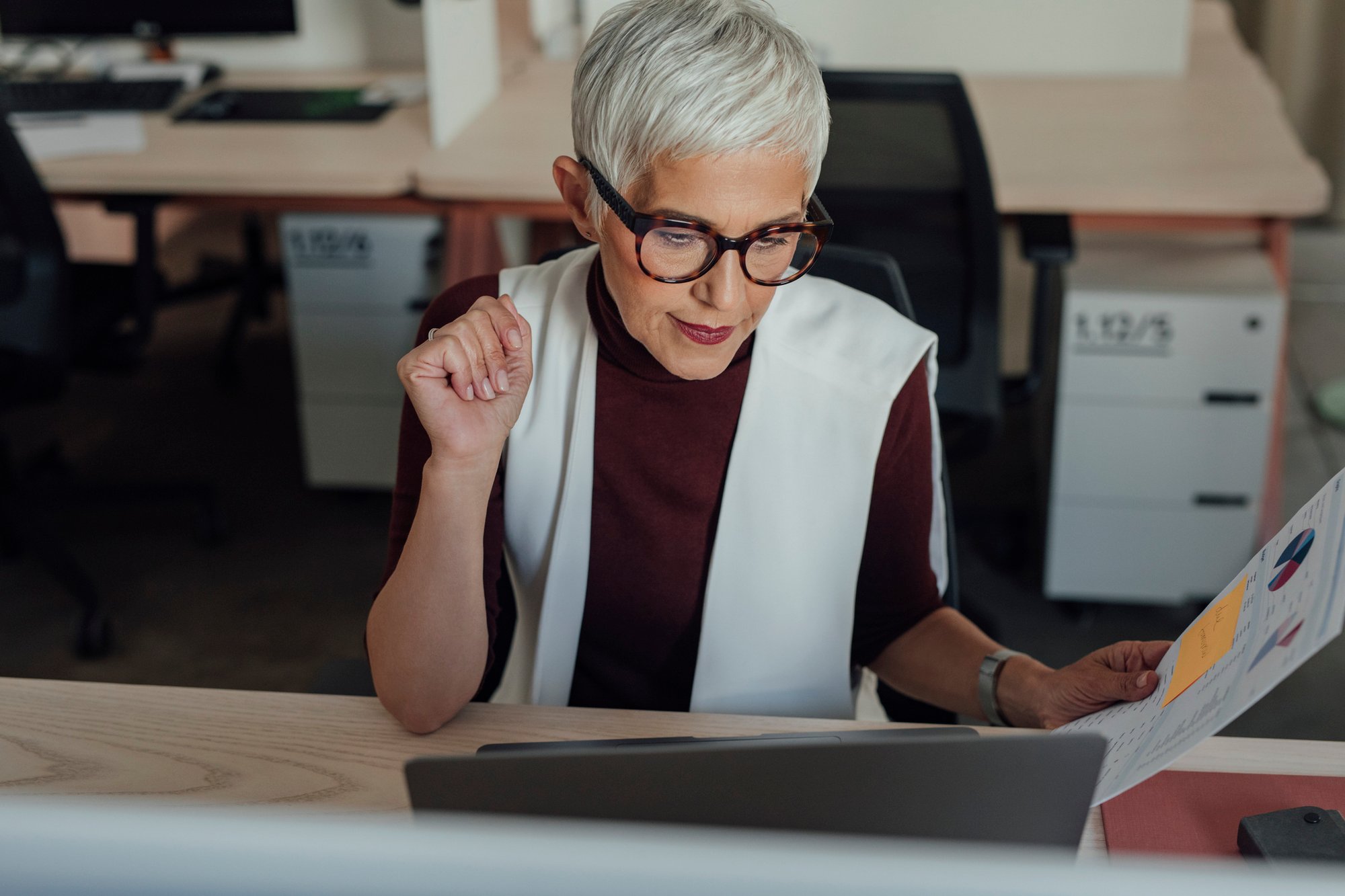 Professional woman with short gray hair and glasses reviewing a document at her desk, focused and engaged in a modern office setting.