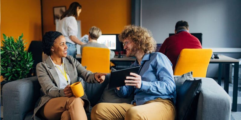 Two coworkers smiling and engaging in a casual discussion on a sofa in a collaborative office space, with others working in the background.