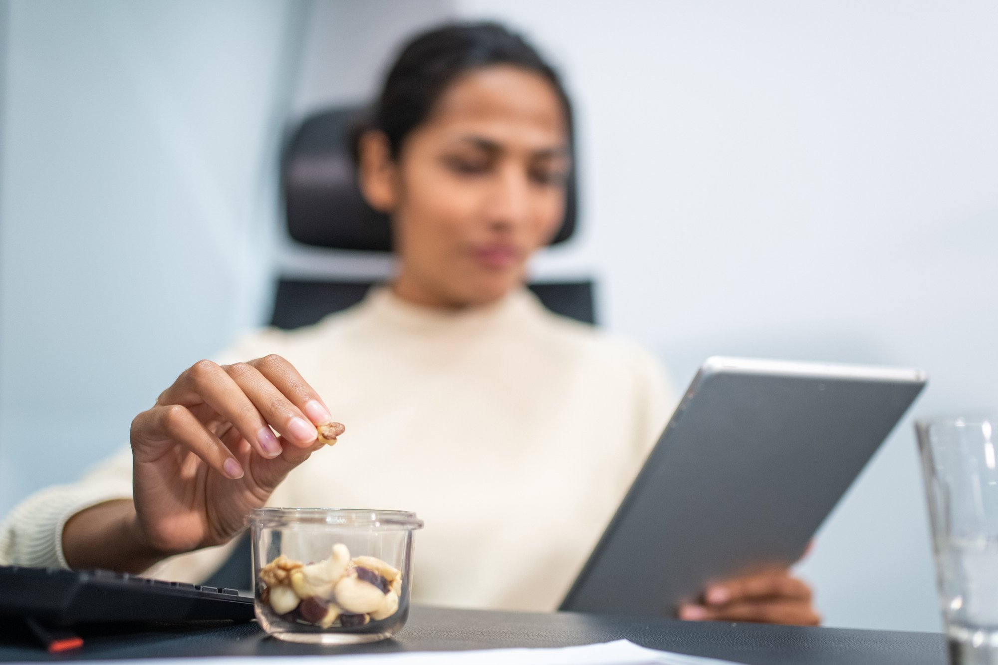 Woman sitting at a desk, focused on a tablet in one hand while picking mixed nuts from a container with the other.