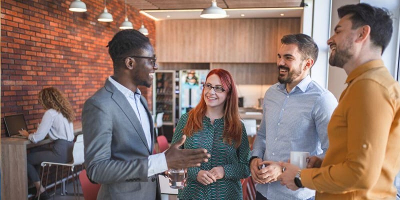 A group of coworkers laughing and having a lively conversation in a modern office break area with a brick wall and cozy ambience.