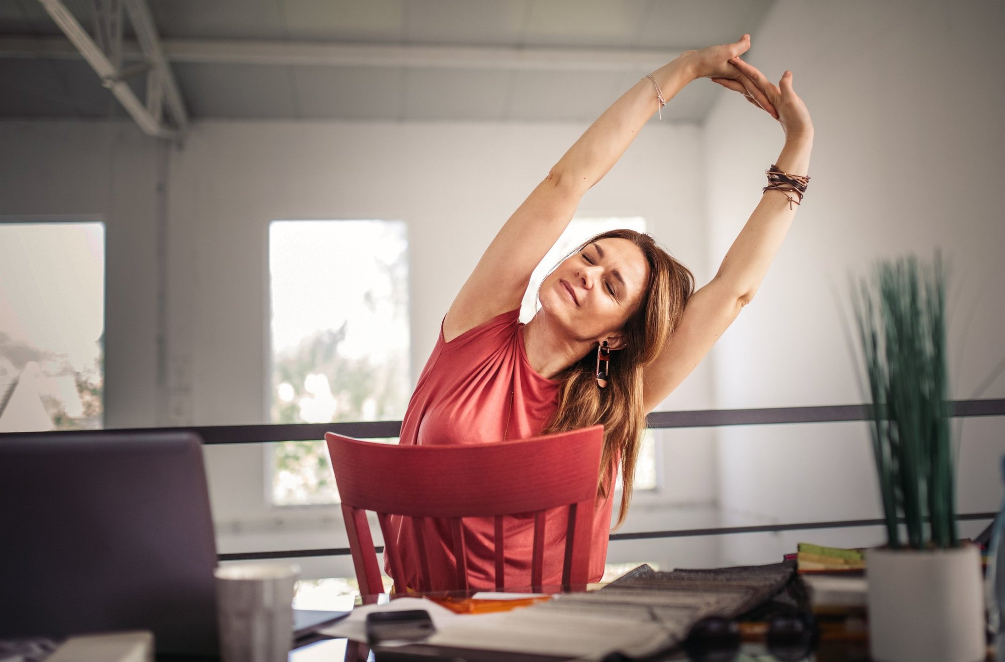 Woman in a red top sitting at a desk, eyes closed, stretching her arms above her head, taking a relaxing break in a bright office space.