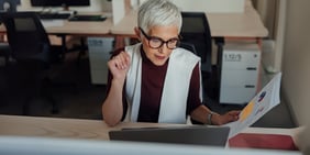 Professional woman with short gray hair and glasses reviewing a document at her desk, focused and engaged in a modern office setting.