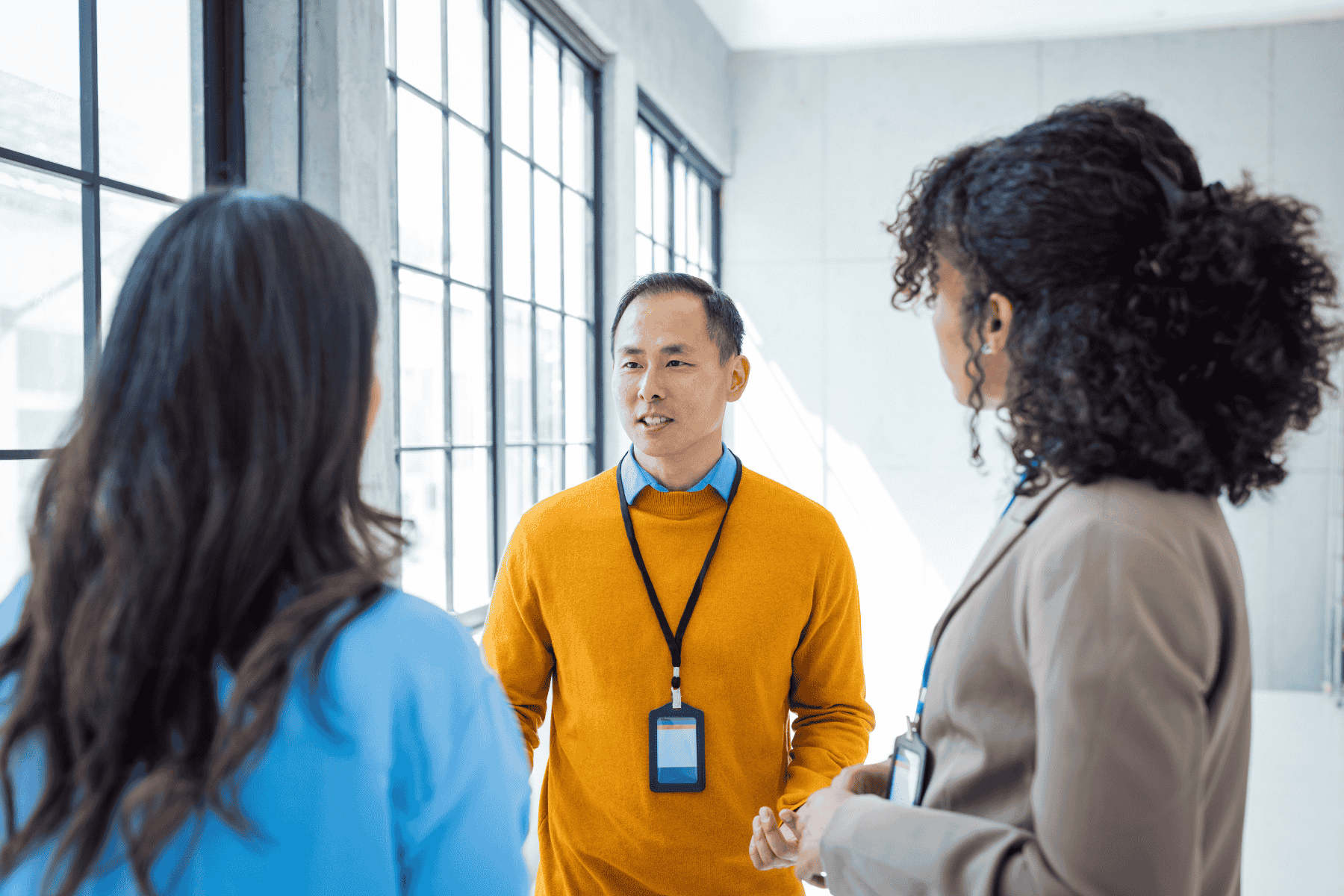  Man in a yellow sweater engaged in a focused discussion with two colleagues in a bright, modern workspace with large windows.