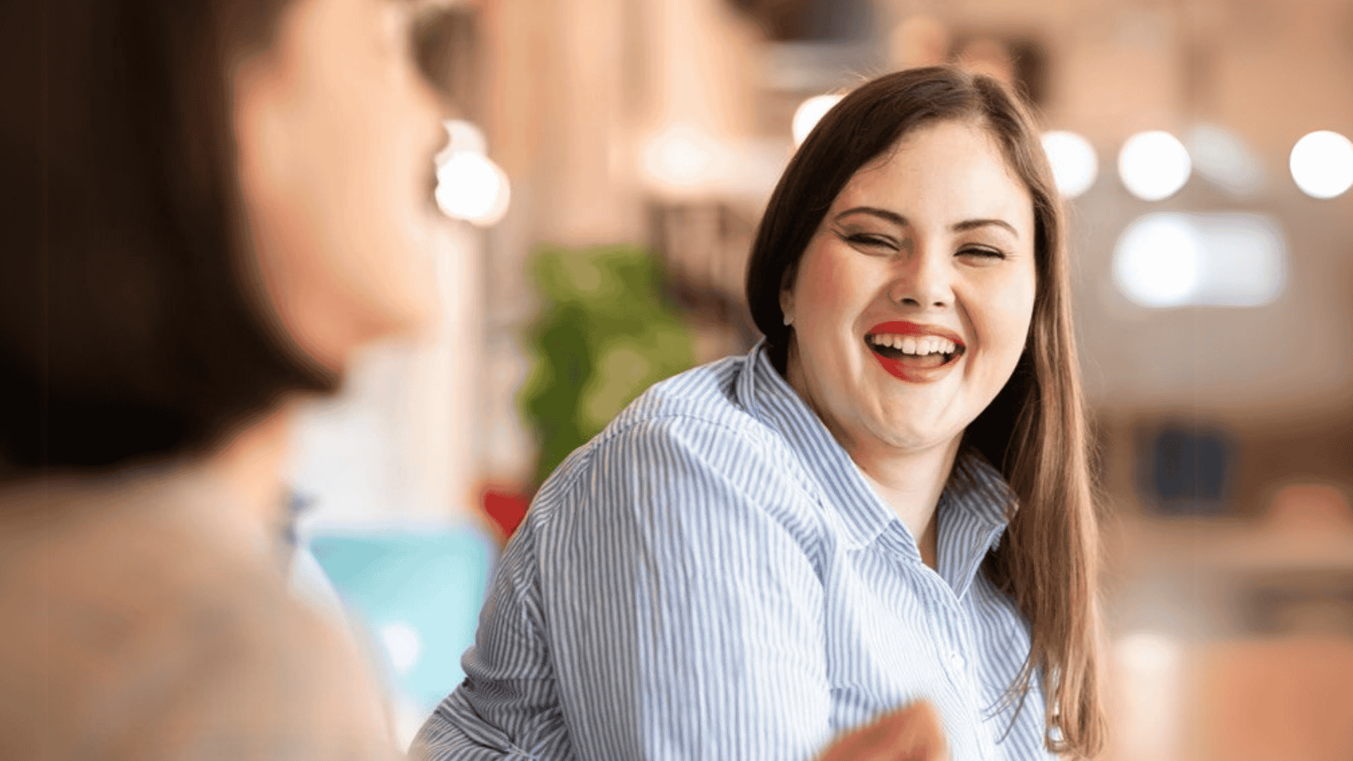  Smiling woman in a blue striped shirt engaged in a joyful conversation in a warm, well-lit office environment.